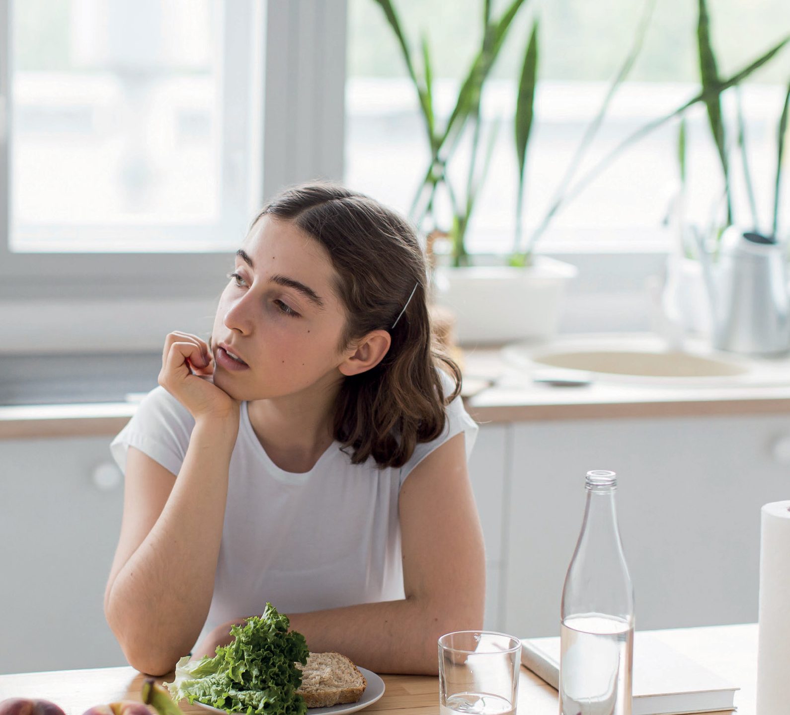 Jovem pensativa apoiando o queixo na mão diante de um prato de salada e frutas na cozinha